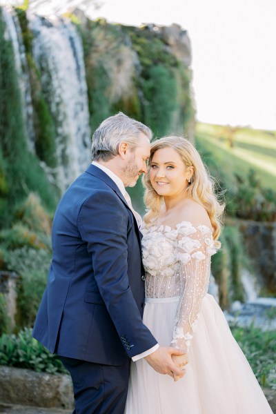 Groom goes in for a kiss as bride looks at camera waterfall in background