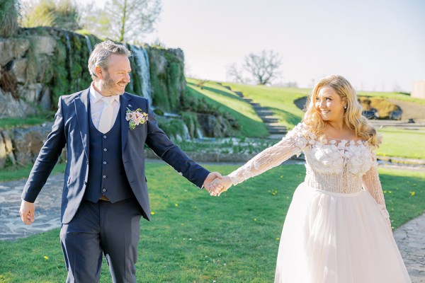 Bride and groom hold hands in garden wedding venue in background they walk on the grass