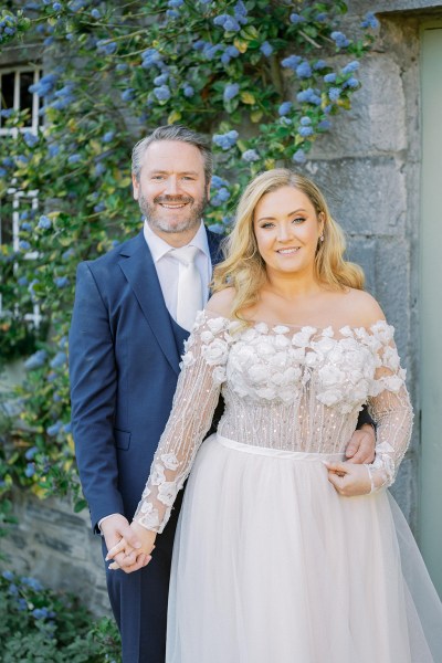 Bride and groom hold hands in garden facing camera