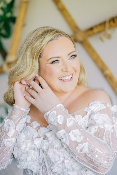 Blonde bride smiles as she puts on earrings