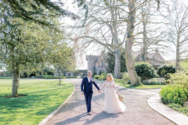 Bride and groom walk smiling holding hands in garden setting wide shot