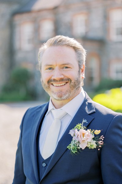 Groom stands in the courtyard smiling flower brooch detail