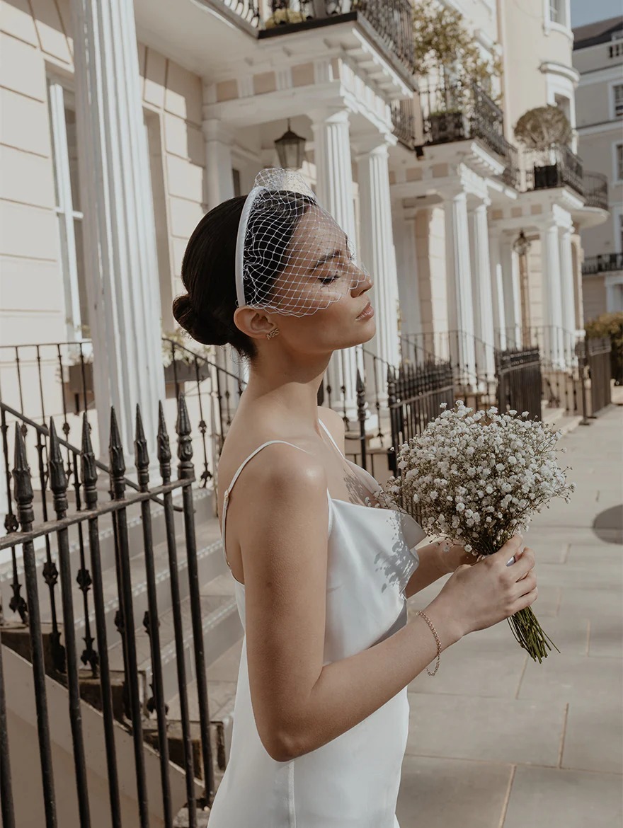 A bride stands on a street in front of a railing and pillars wearing a satin wedding dress and a birdcage veil while holding a bouquet of white flowers