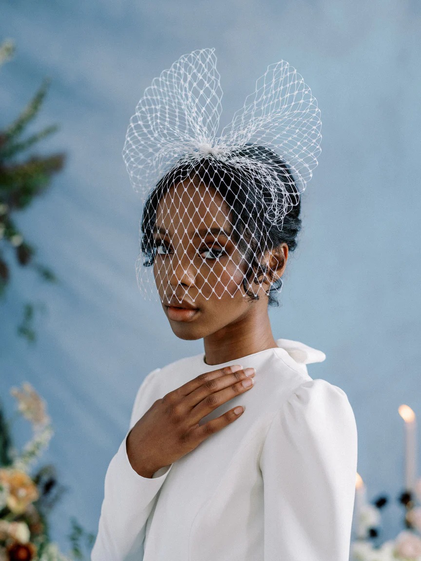 A bride wears a bow birdcage veil with a white dress holding her hand to her neck standing in front of a blue background