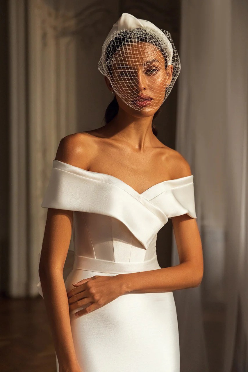 A bride stands in front of a cream curtain wearing an off-the-shoulder dress and a headband with a birdcage veil