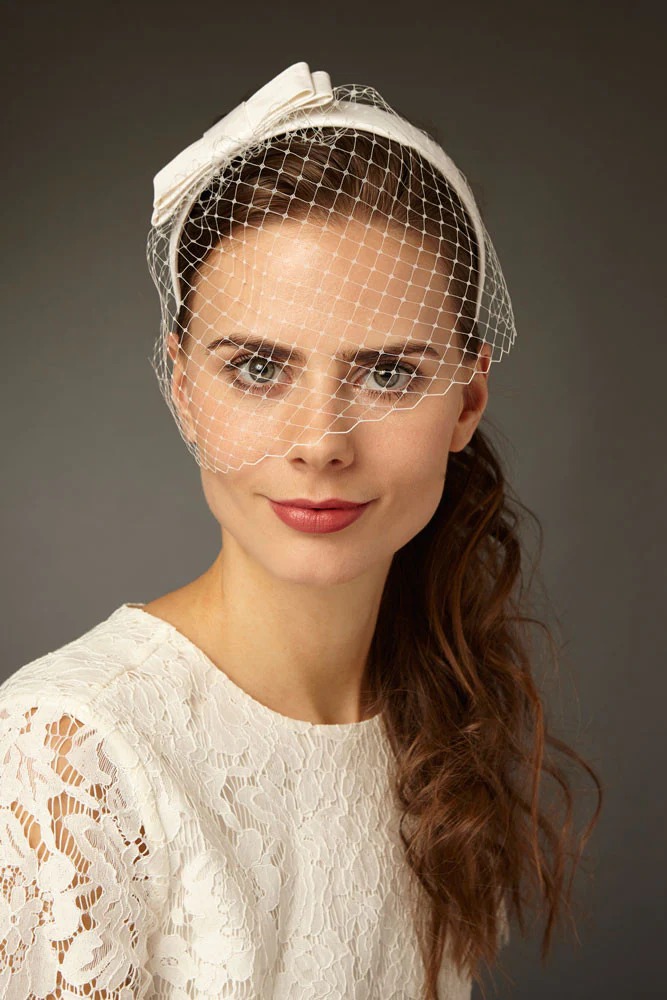 A female model seen against a dark grey background wearing a lace dress and a bow headband with a netted cover