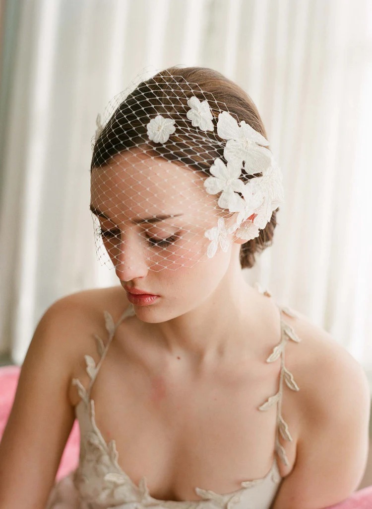 A female model wears a lace wedding dress with a veil with floral accents sitting in front of a white curtain