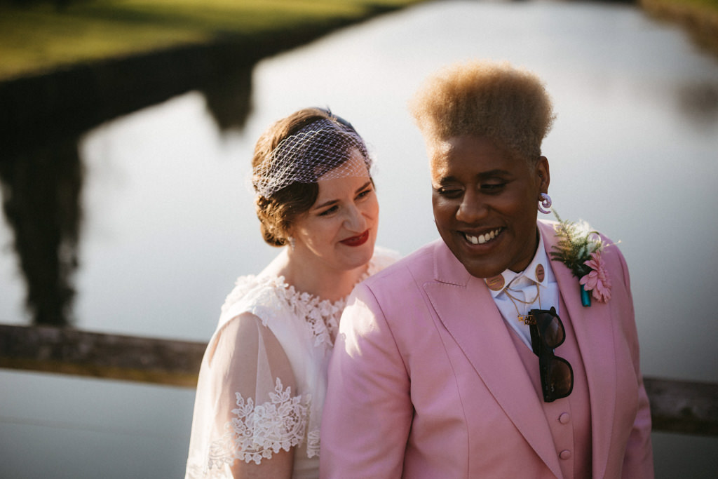 Two brides one in dress and one in pink suit smile lake in background