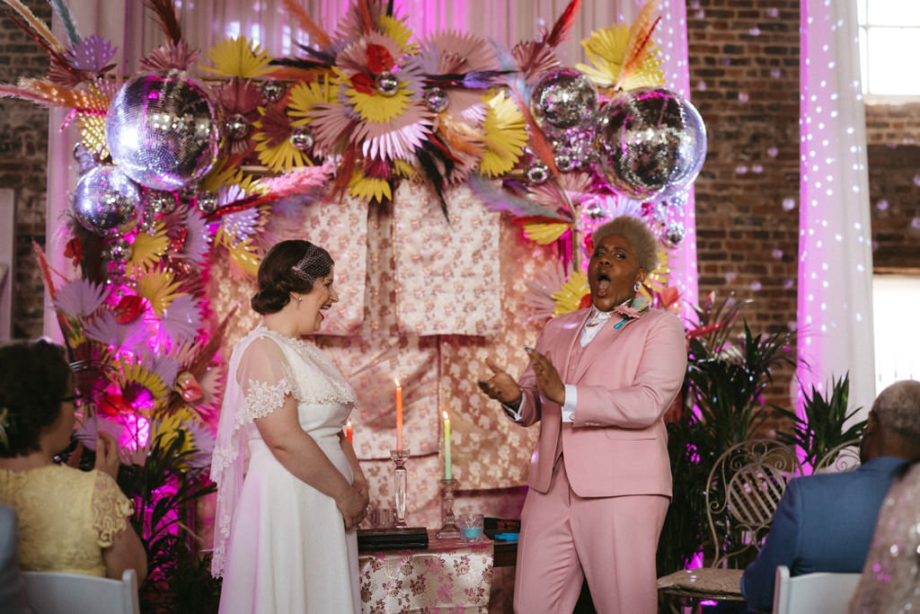 Two brides clap and laugh at alter disco ball colourful setting