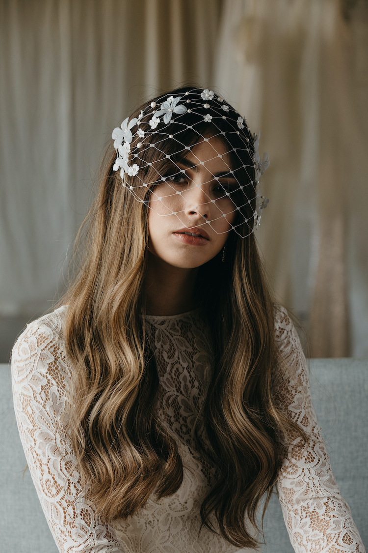 A female model sits on a couch wearing a lace wedding dress and a birdcage veil with floral accents