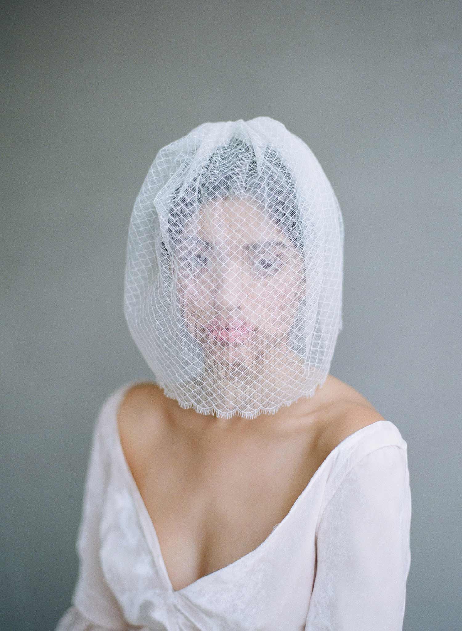 A bride sits against a dark grey background wearing a white wedding dress with a birdcage veil