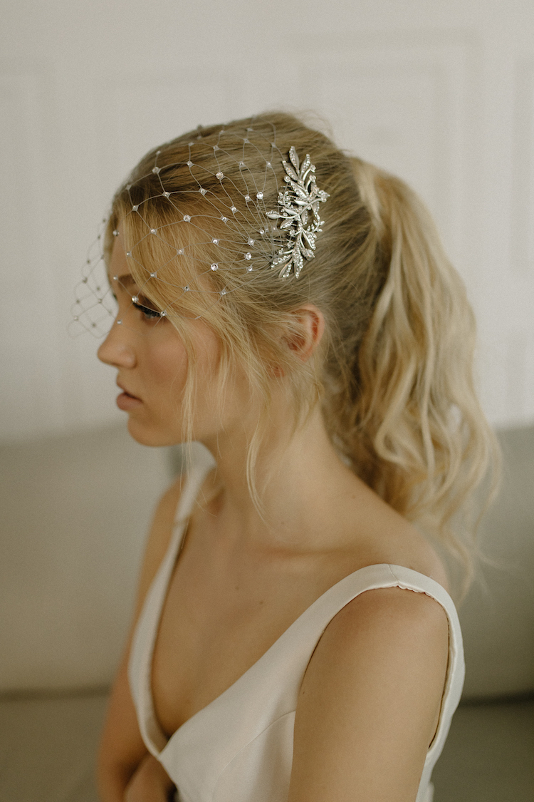 A bride sits on a cream couch wearing a crystal netted headpiece and a white dress