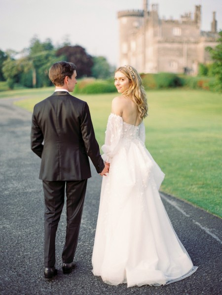 Bride and groom walk along the pathway to wedding venue bride looks over her shoulder