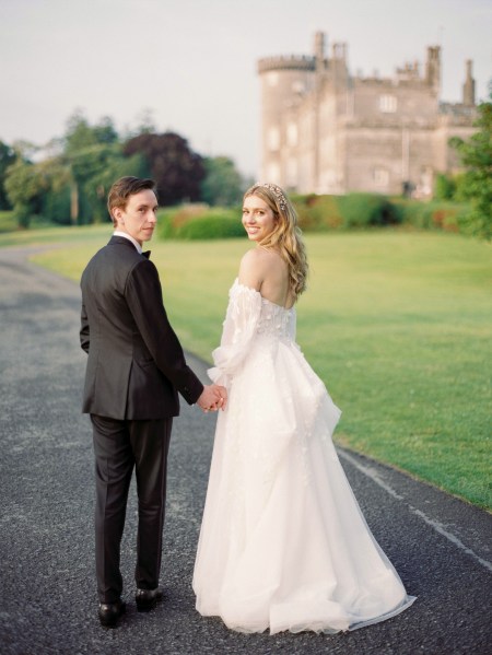 Bride and groom walk along the pathway to wedding venue both look over their shoulders