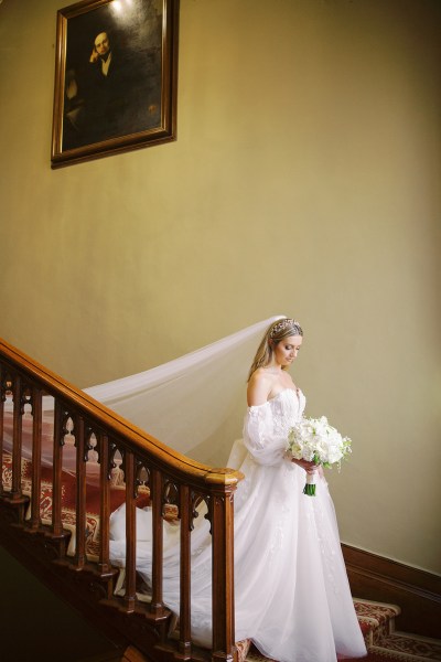 Bride walks down the staircase train and veil following she holds a white rose bouquet