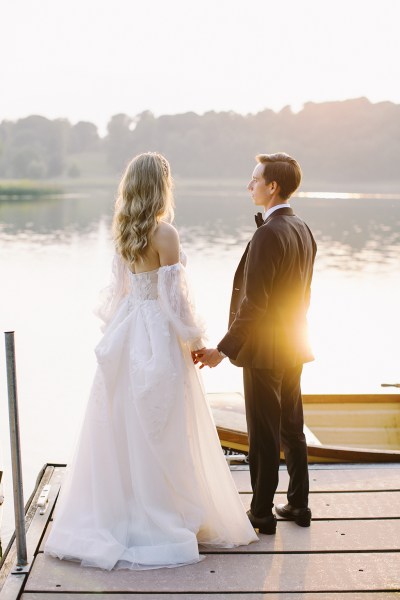 Bride and groom stand on boardwalk in front of lake sun is setting boat