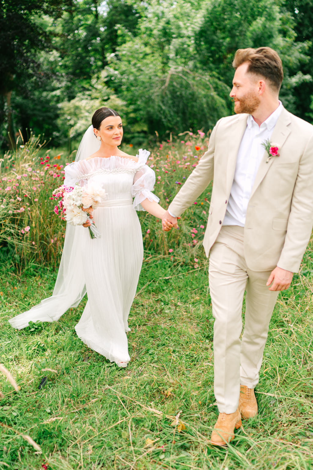 Bride and groom walk hand in hand on the grass looking at each other