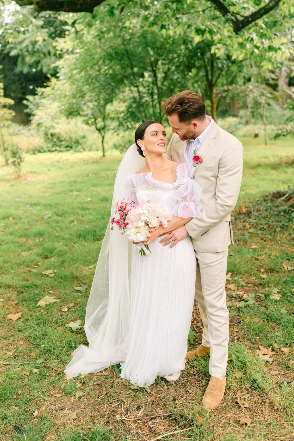 Bride and groom stand in garden park and smile at each other standing on grass