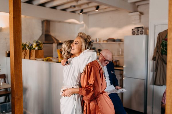 Bride wearing white satin robe hugs a bridesmaid wearing a burnt orange wedding robe