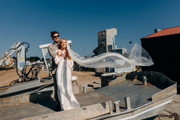 A bride and groom stand on an old boat with her veil flowing through the air