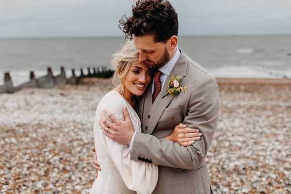 Bride and groom face each other on the beach sand and rocks sea in background