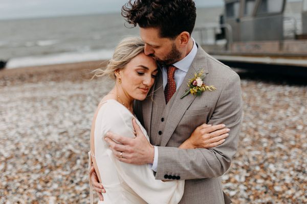 Bride and groom smile at each other on beach setting sand and rocks sea in background kiss on the head