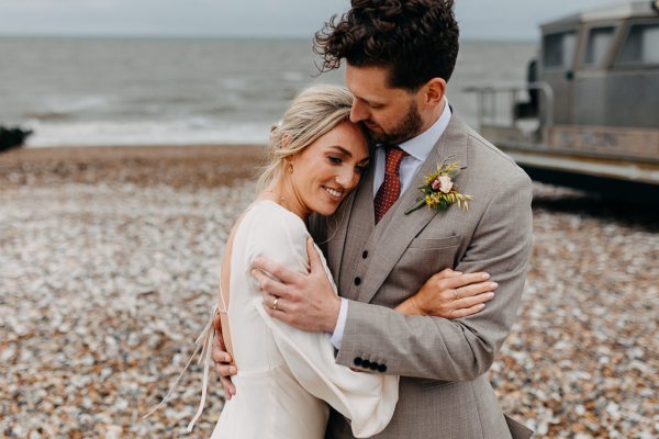 Bride and groom smile at each other on beach setting sand and rocks sea in background kiss on the head