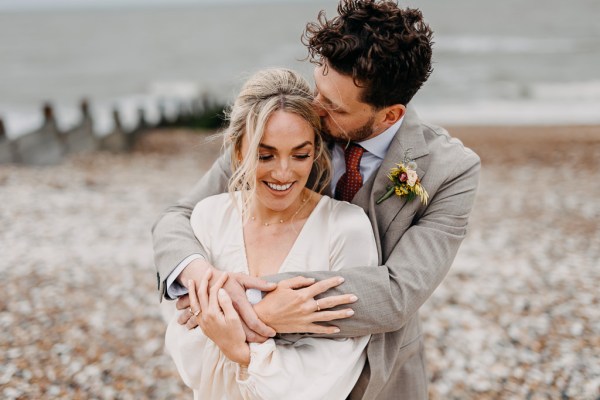 Groom hugs bride from behind on beach setting sand and rocks sea in background kiss on the head