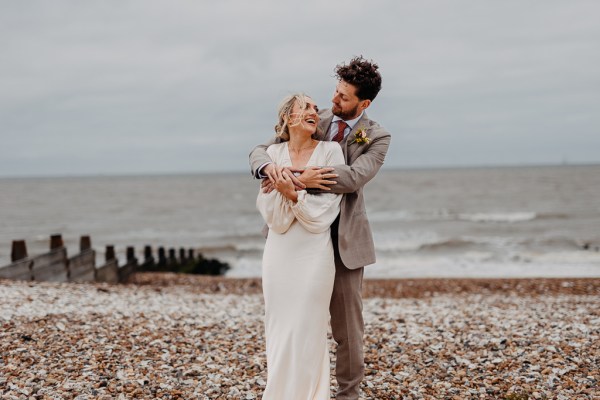 Groom hugs bride from behind on beach setting sand and rocks sea in background