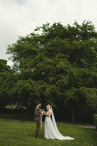 Bride and groom stand in the garden together trees in background