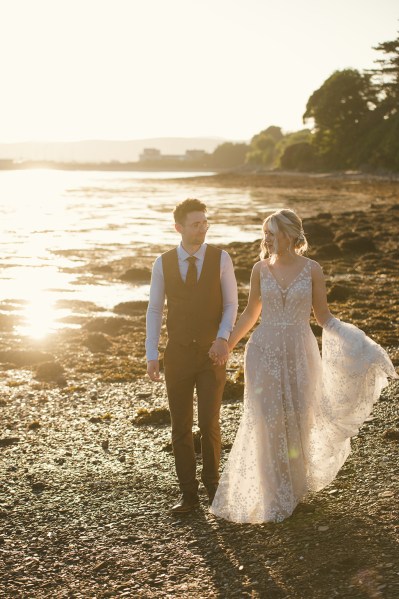 Bride and groom in beach setting sunset behind couple they walk along the sand