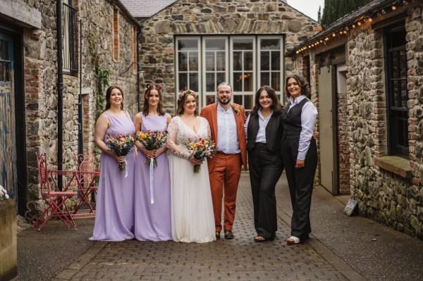 Groom in orange bride and bridesmaids in purple dresses groomsmen all smiling exterior shot