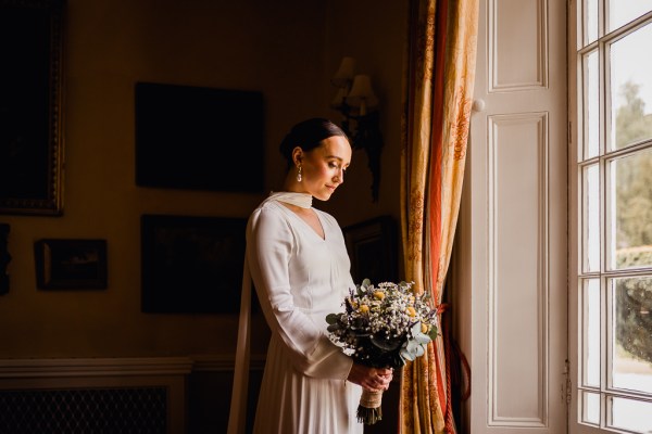 Bride looks down at flowers/bouquet stands beside window