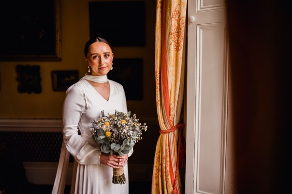 Bride faces camera at flowers/bouquet stands beside window