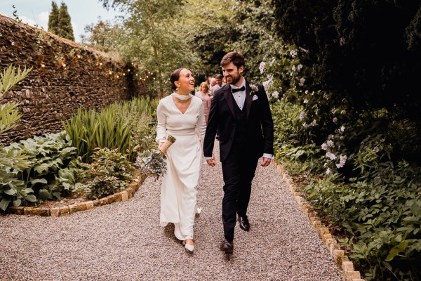 Bride and groom smile walking down pathway to garden