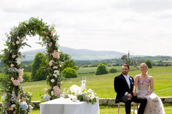 Bride and groom seated during ceremony wide shot of background grass mountains and flower bed arch and guests