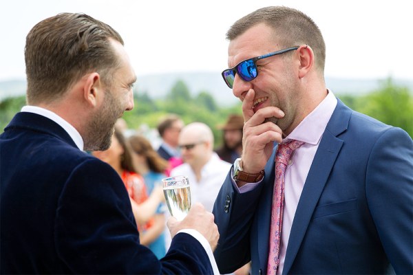 Two men one wearing sunglasses has hand over mouth smiling