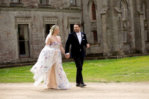 Bride and groom hold hands on the grass in front of wedding venue wide shot