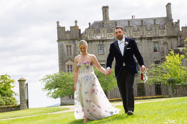 Bride and groom hold hands on the grass in front of wedding venue wide shot