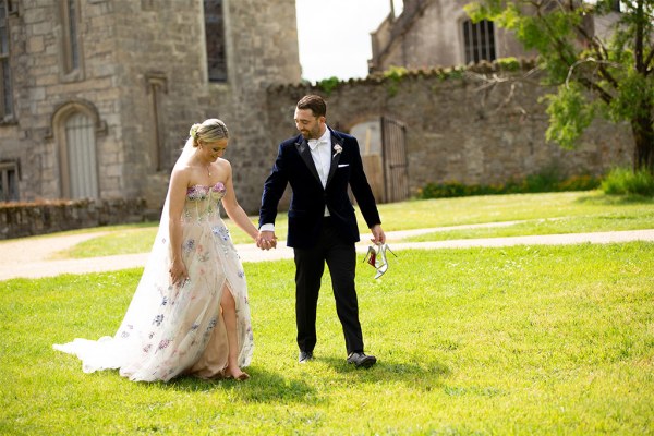 Bride and groom hold hands on the grass in front of wedding venue wide shot