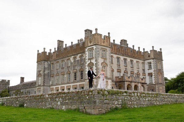 Bride and groom stand on ledge balcony to wedding venue wide shot of castle