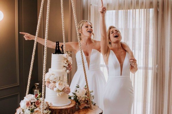 Brides cheer on crowd beside white wedding cake