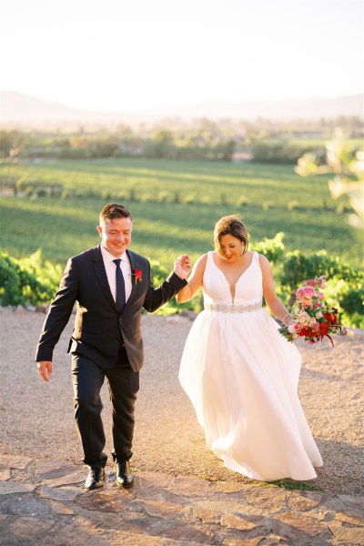 Bride and groom walking uphill grass and farm in background to couple