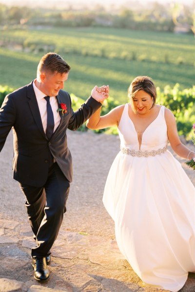 Bride and groom walking uphill grass and farm in background to couple