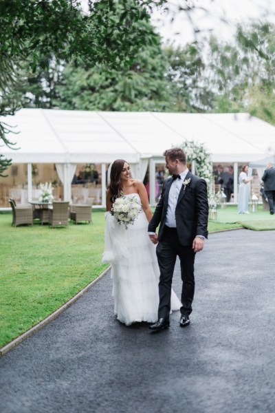 bride and groom look at each other as they walk the pathway marquee tent in background