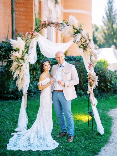 Bride and groom stand in front of flower bed holding glasses of prosecco