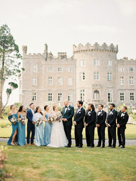 bride groom groomsmen and bridesmaids stand on grass in front of castle setting