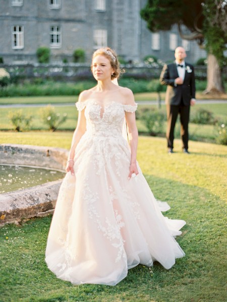 bride stands in front of groom and waits both on grass fountain in shot