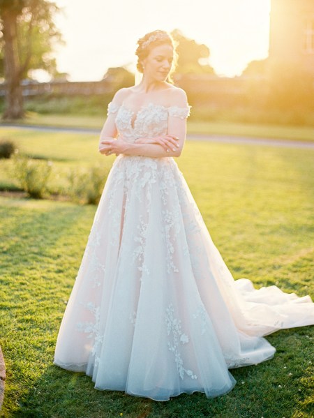 bride stands on her own on green grass sun is setting behind her