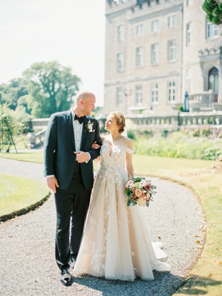 bride and groom hold hands on pathway to wedding castle venue sun is shining